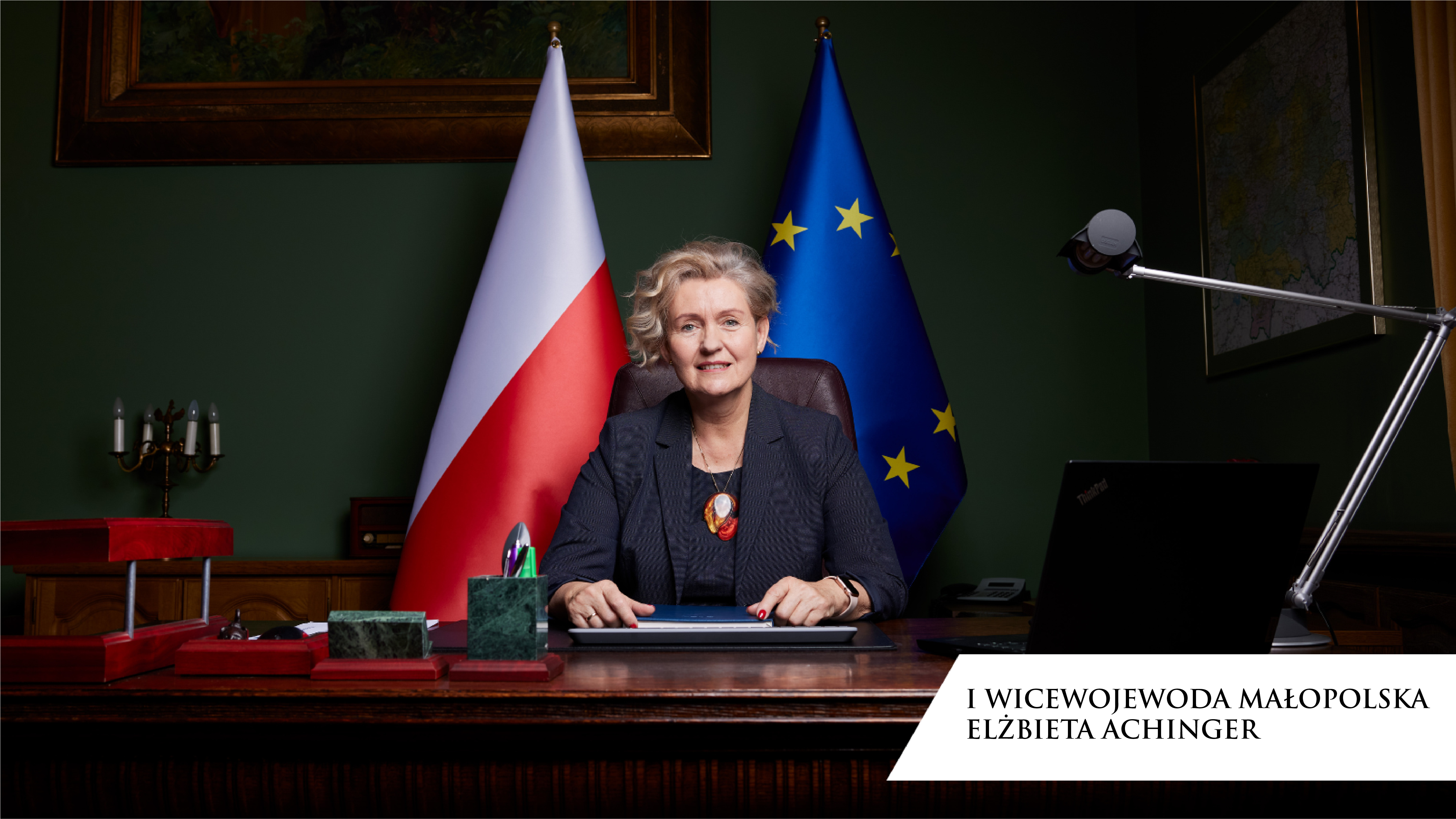 Elżbieta Achinger, I Wicewojewoda Małopolska siedząca przy biurku na tle flag Polski